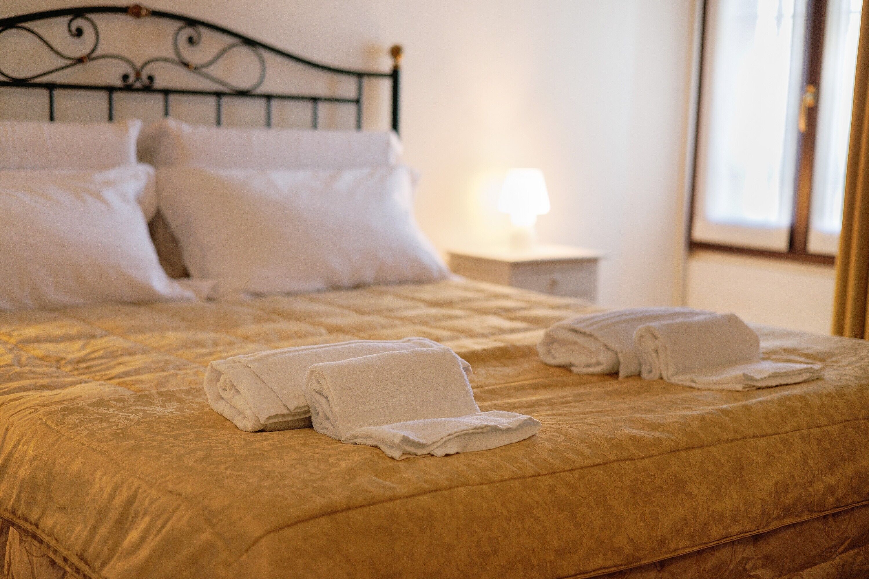 Una cama bien hecha con una colcha dorada y almohadas blancas, una mesita de noche con una lámpara, y una ventana con cortinas.