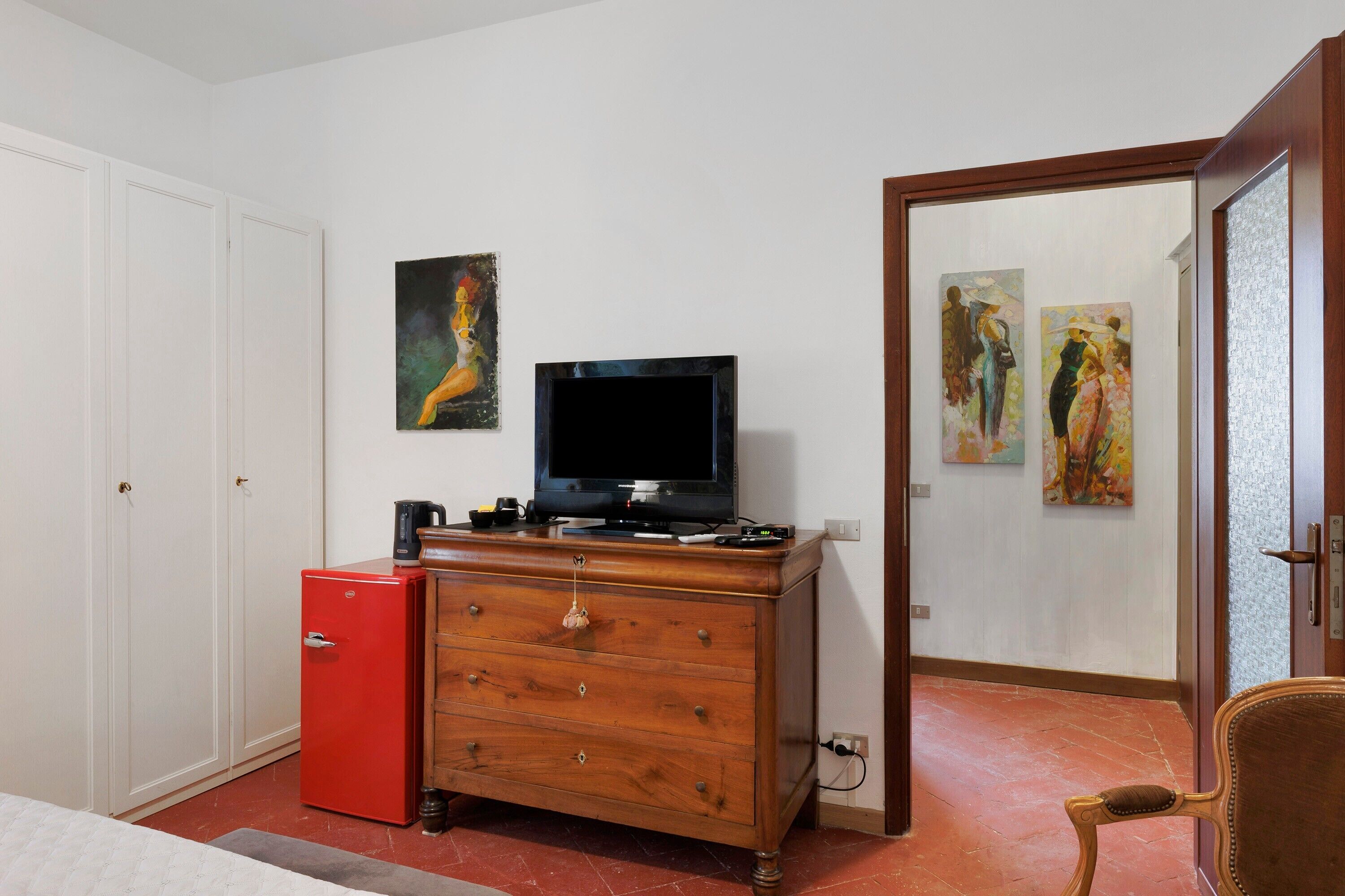 Una habitación con un televisor sobre un aparador de madera, un refrigerador rojo y dos cuadros en la pared.