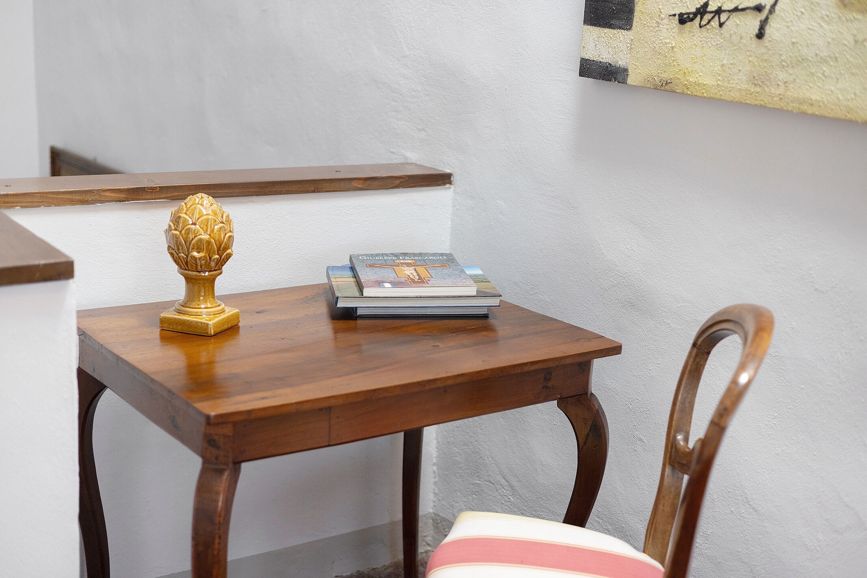 A small wooden table with a decorative item and a book, a wooden chair, and a white wall with a painting.