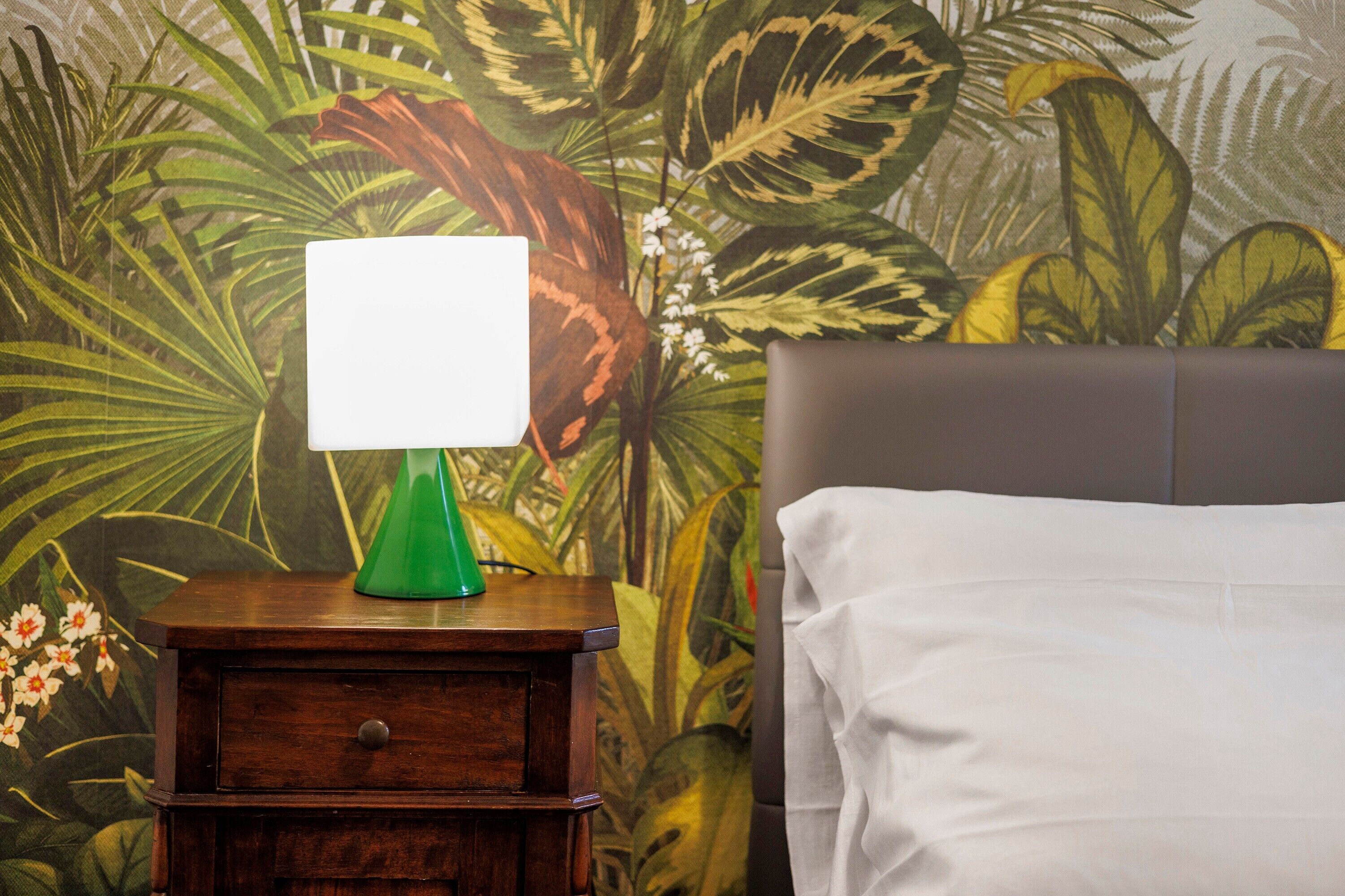Un lit avec des draps blancs, une lampe verte posée sur une table de chevet en bois et un papier peint aux motifs tropicaux.