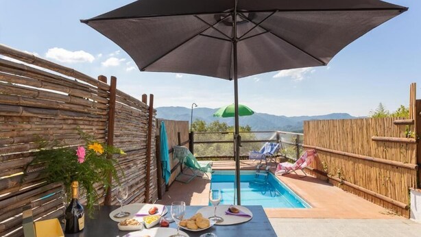 Un área de comedor al aire libre con una mesa para dos, una botella de champán y vistas a una piscina y montañas al fondo.