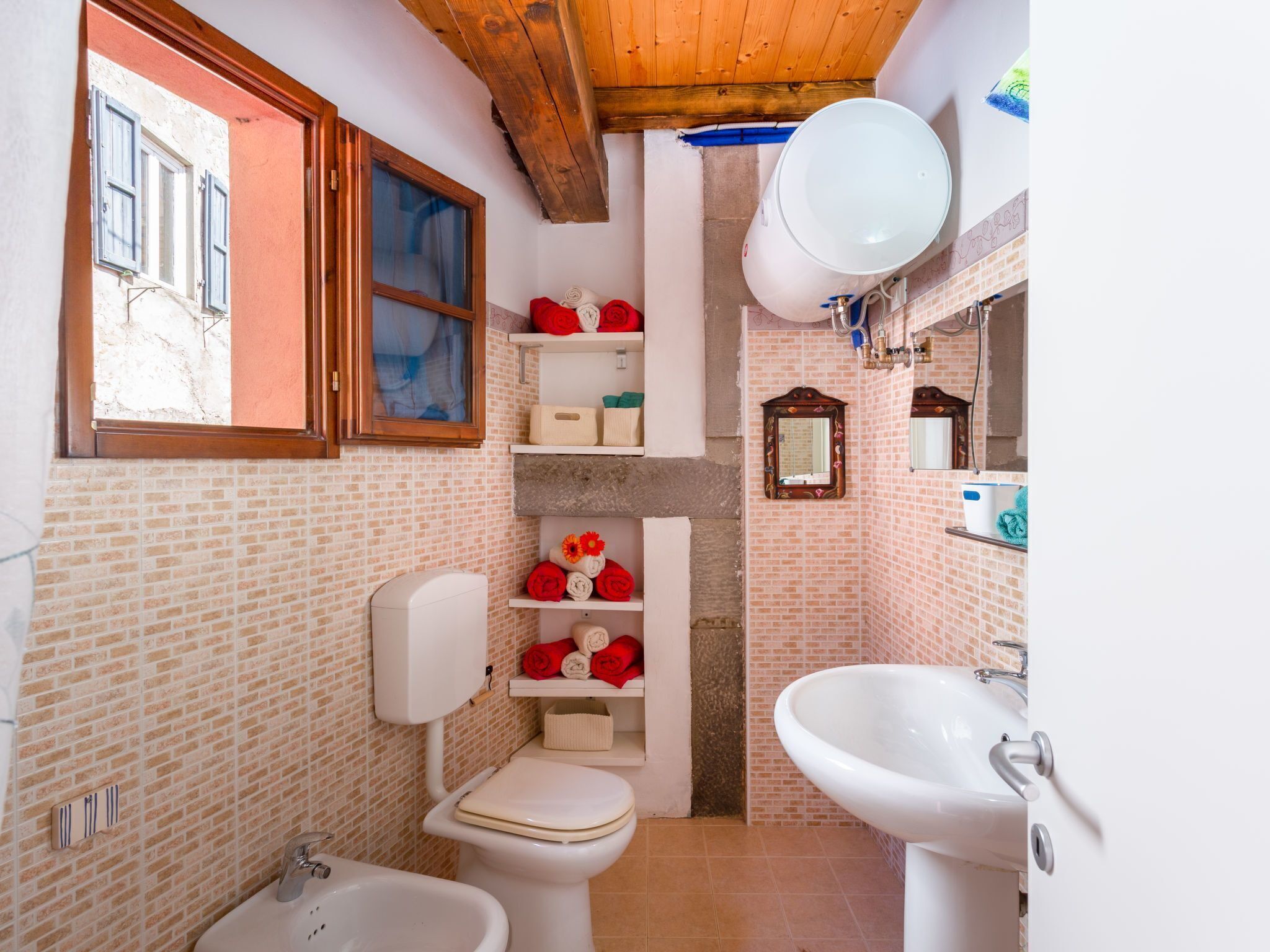 A bathroom with a toilet, bidet, and sink, a large water heater, and a window with shutters.