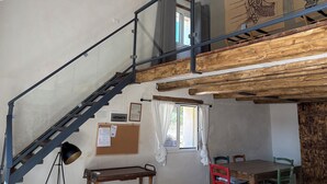 Una habitación con escalera de madera, una mesa, sillas, una ventana con cortinas y un tablón de corcho con papeles.