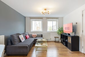 Ein modernes Wohnzimmer mit einem grauen Sofa, einem Glastisch, einem Fernseher, der 'GUEST READY' anzeigt, und einer Topfpflanze.