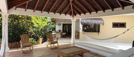 A covered outdoor seating area with a hammock, wooden chairs, and a sofa.