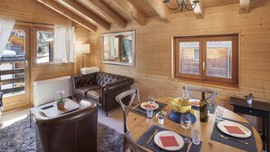 Interior acogedor de una cabaña de madera con una mesa de comedor para dos personas, un sofá de cuero y ventanas amplias.