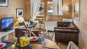 Interior de una cabaña de madera con televisor, sofá de cuero, mesa de comedor y vista a un paisaje nevado a través de la puerta.
