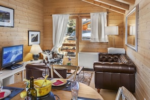 L’intérieur d’un chalet en bois, avec un téléviseur, un canapé en cuir, une table à manger et une vue sur un paysage enneigé par la porte.