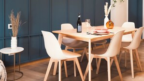 Uma área de refeições com mesa de madeira, cadeiras brancas, uma mesinha lateral com um vaso e um relógio na parede.