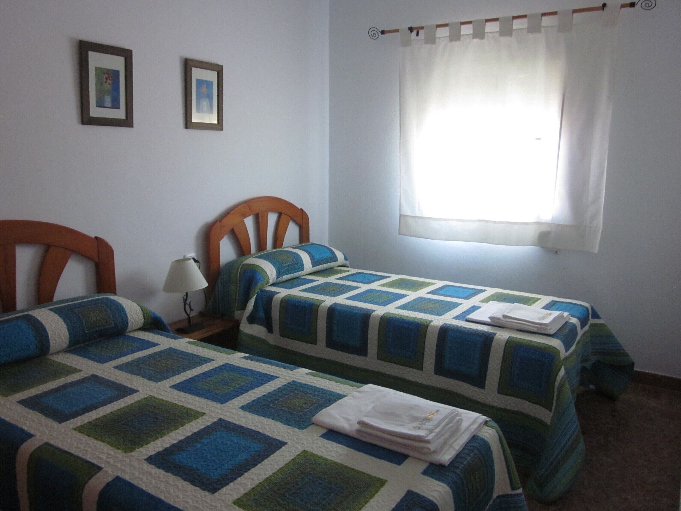 Ein Zimmer mit zwei Einzelbetten, einem Nachttisch mit Lampe und einem Fenster mit weißem Vorhang.