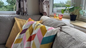 En sofa med farverige puder, et vindue med gardiner og en potteplante på et vindueskar.