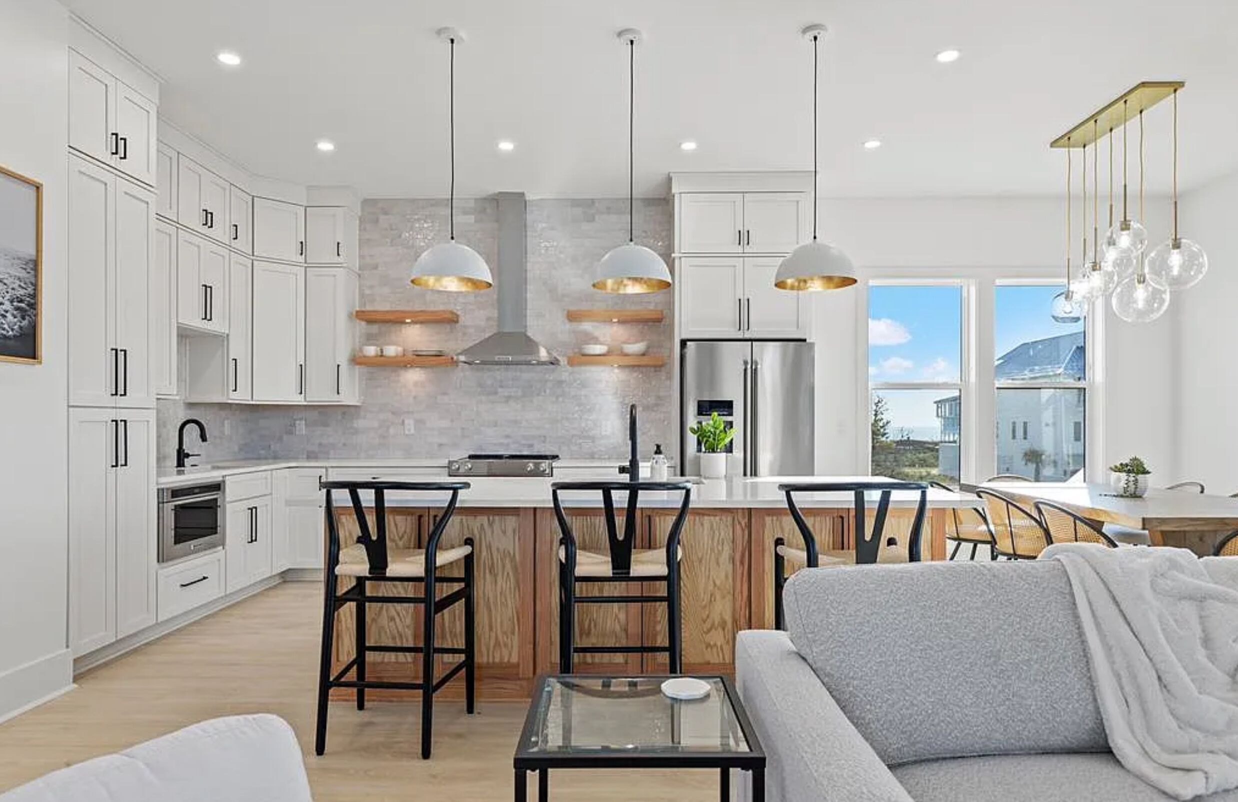 Une cuisine moderne avec des meubles blancs, un îlot central, des luminaires suspendus et une vue sur l’extérieur.