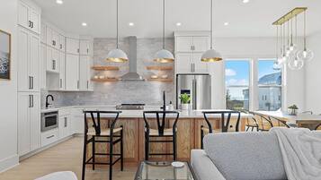 Une cuisine moderne avec des meubles blancs, un îlot central, des luminaires suspendus et une vue sur l’extérieur.
