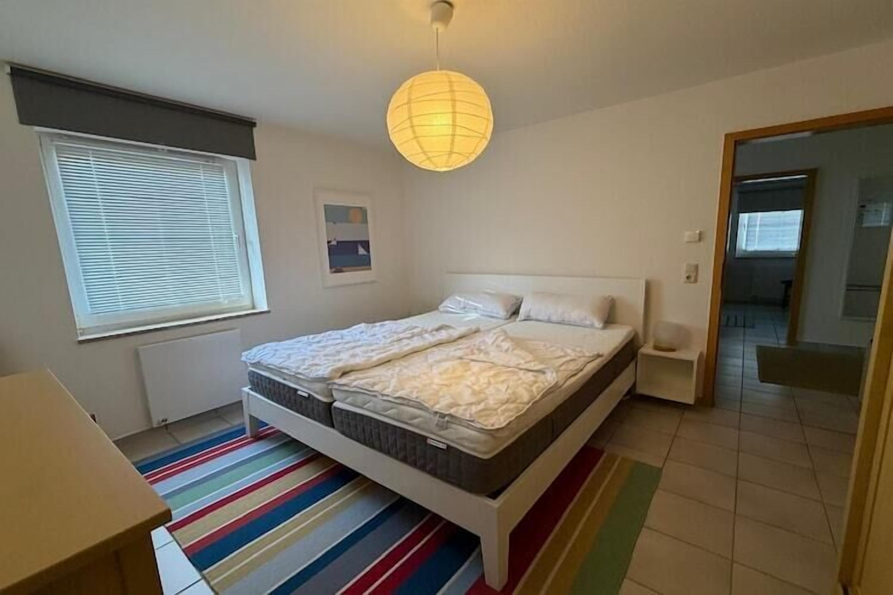A bedroom with a bed, a bedside table, a window with blinds, a radiator, a colorful striped rug, and a hanging light fixture.