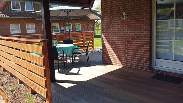 Une terrasse extérieure avec des bancs en bois et une table, un mur en briques et une porte dotée d’un panneau de verre.