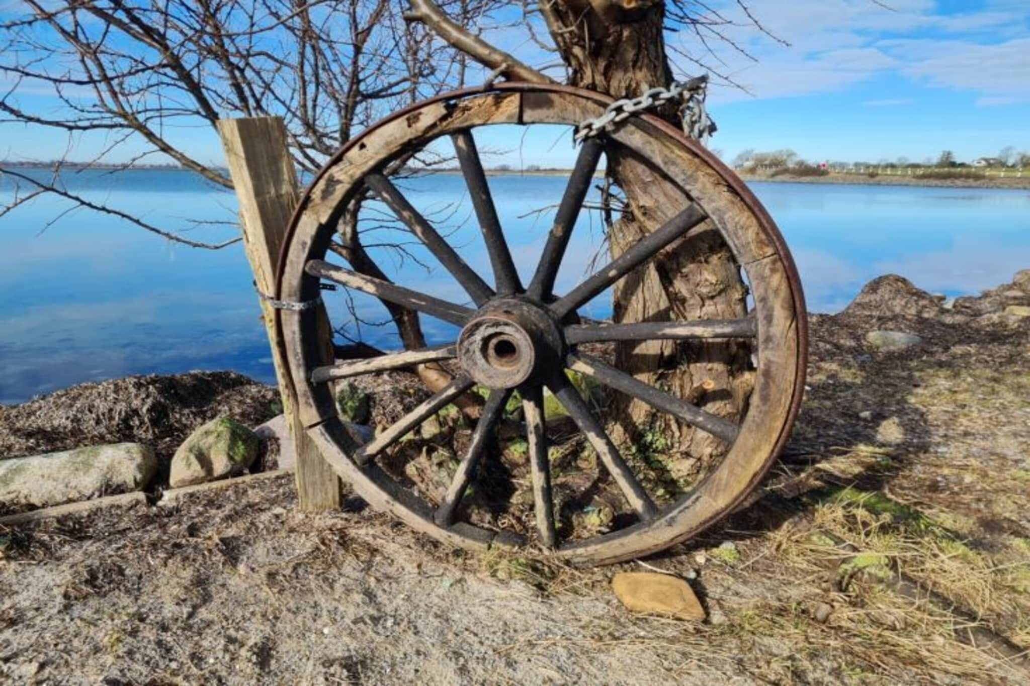 A large, rusty wooden wheel with a chain, positioned against a backdrop of a calm lake and bare trees.