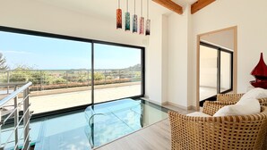A modern interior with a large glass window, a glass pool, wicker furniture, and hanging pendant lights. - Peaceful Villa with Pool, Jacuzzi & Gym (Antibes)