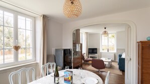Ein Essbereich mit Tisch für zwei Personen, einer Weinflasche und einem Weinglas. Im Hintergrund befindet sich ein Fernseher, ein Sofa und ein Schrank.