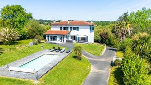 Une grande maison avec une piscine, entourée d’une végétation luxuriante et accessible par une allée sinueuse.