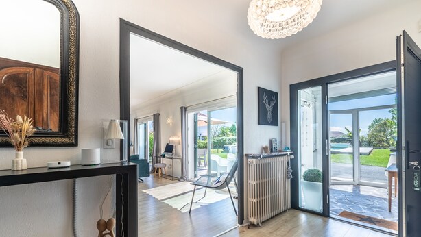 Un intérieur moderne agrémenté d’un grand miroir, d’un lustre et d’une vue sur un espace extérieur accessible par des portes vitrées.