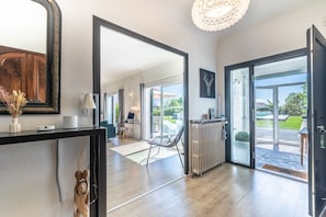 Un intérieur moderne agrémenté d’un grand miroir, d’un lustre et d’une vue sur un espace extérieur accessible par des portes vitrées.