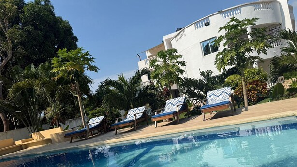 A swimming pool with lounge chairs, surrounded by tropical vegetation and a white building with balconies. - cosy by-beach retreat!Warm private escape with private pool (Kwale County)
