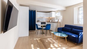 A modern living room with a flat-screen TV, a blue sofa, a glass coffee table, and a kitchen area with white cabinets and a marble countertop. - Le Cocoon - Avec jardin de 900m2 (La Celle-Saint-Cloud)