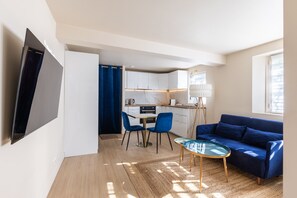 A modern living room with a flat-screen TV, a blue sofa, a glass coffee table, and a kitchen area with white cabinets and a marble countertop.