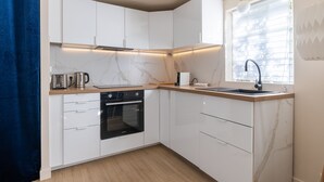 A modern kitchen with white cabinets, a built-in oven, a sink, and a window with blinds. - Le Cocoon - Avec jardin de 900m2 (La Celle-Saint-Cloud)