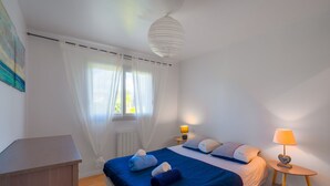 A bedroom with a bed, a bedside table with a lamp, a radiator, and a window with curtains. - Fenêtre sur mer - À 400m de la plage (Saint-Nic)