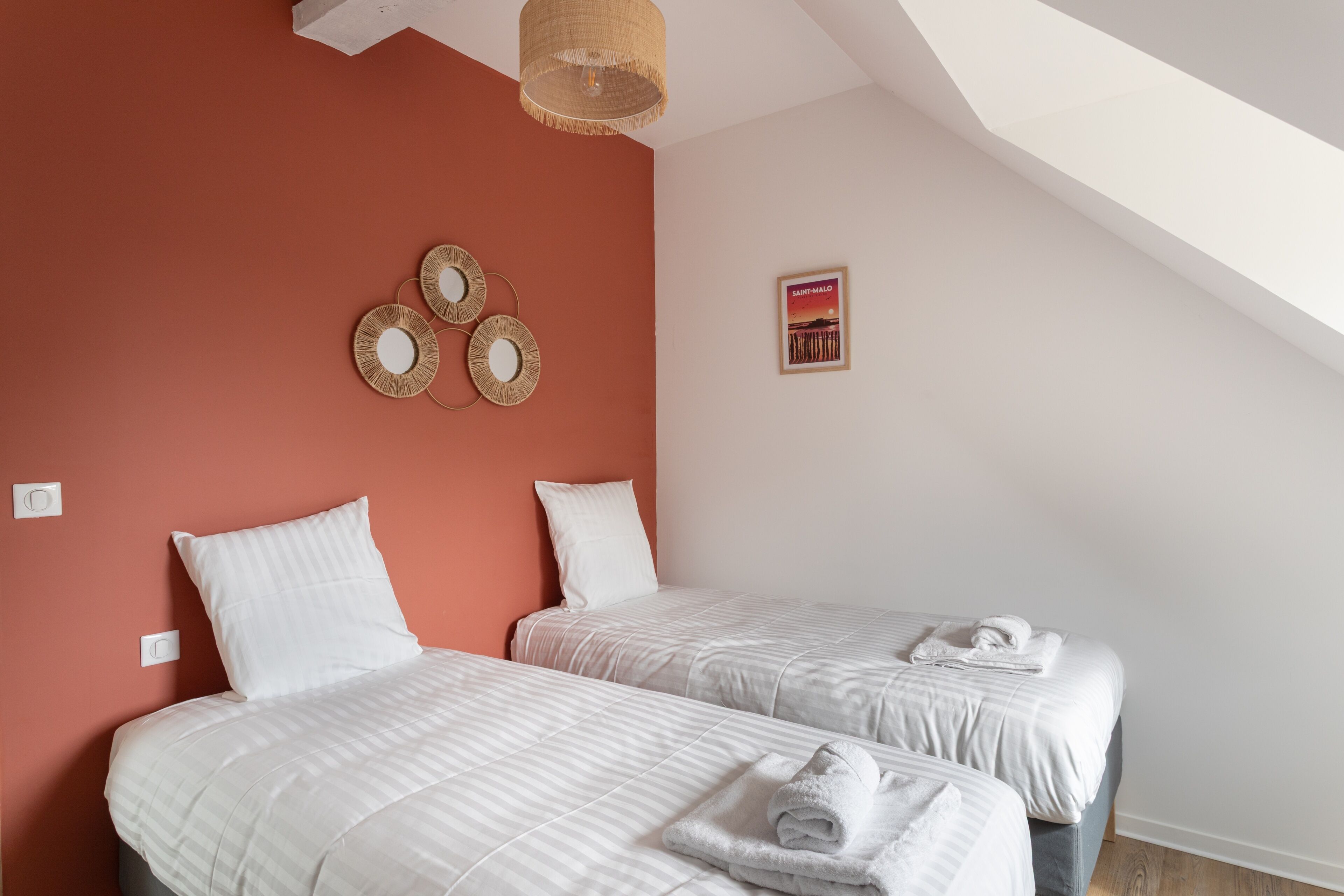 Une chambre avec deux lits, un luminaire au plafond, un miroir fixé au mur et un tableau encadré.