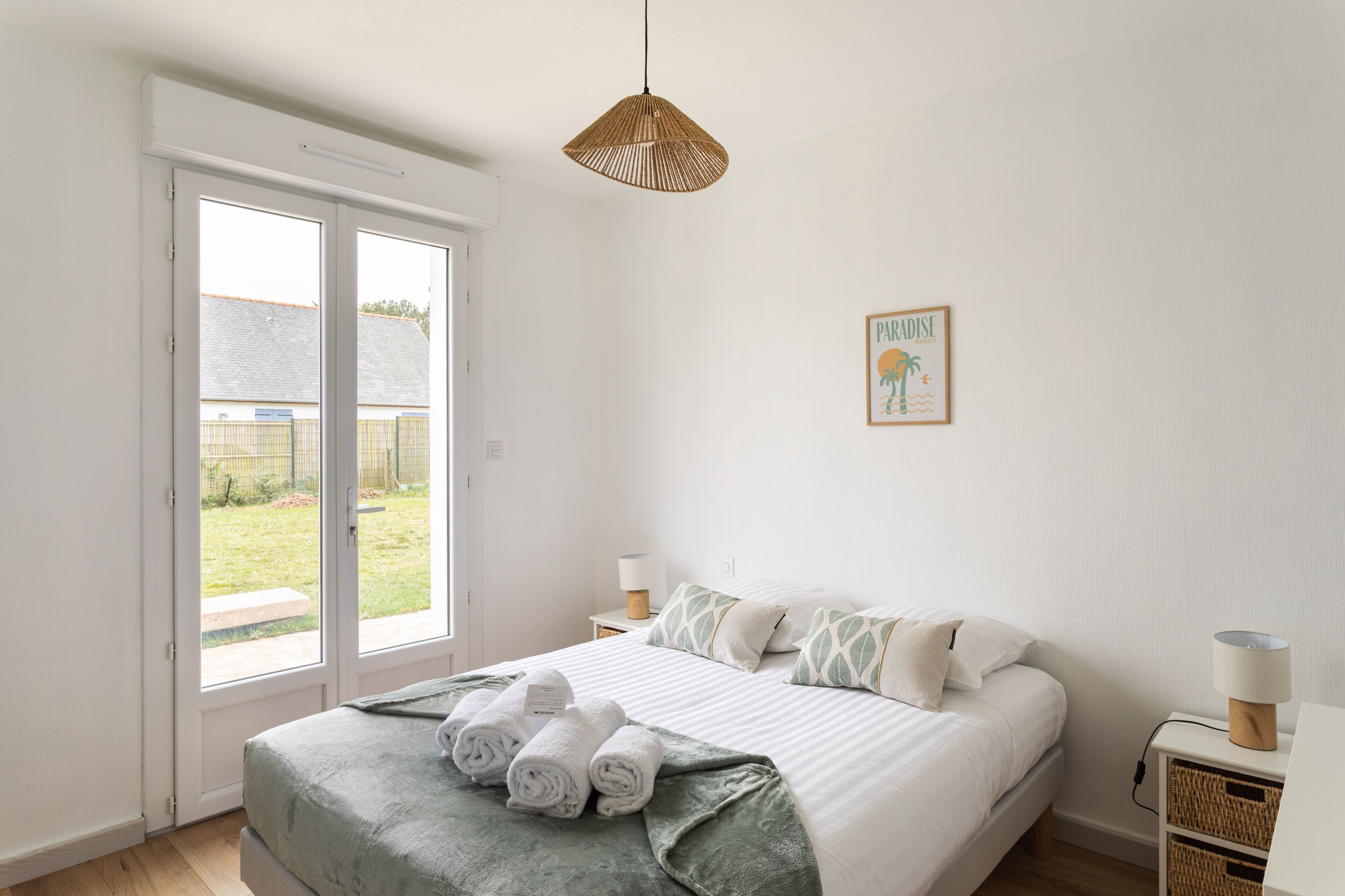 A bedroom with a bed, two bedside tables, a hanging light, a door to a balcony, and a wall-mounted picture.
