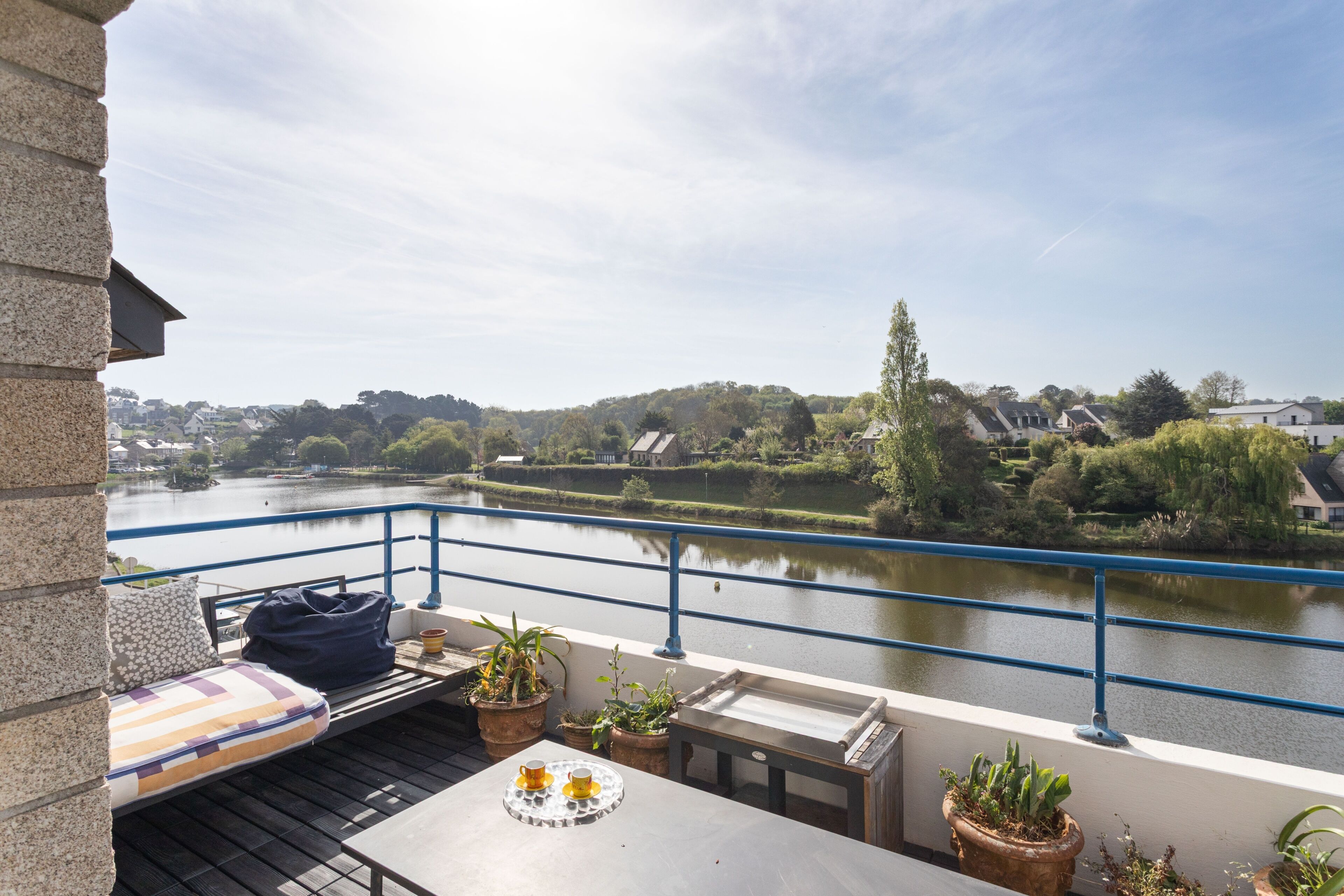 A balcony with a view of a river, potted plants, and a table with a tray of yellow cups.