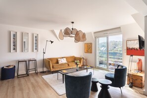 A modern living room with a sofa, coffee table, and chairs, a large window with a view, and a ceiling light fixture. - La Dahouëtine - Blacon Vue sur Étang (Pléneuf-Val-André)