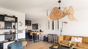 En moderne stue med sofa, sofabord og en blå lænestol. Der er et køkkenområde med spisebord og stole.