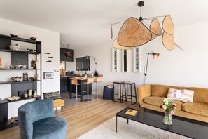 A modern living room with a sofa, a coffee table, and a blue armchair. There is a kitchen area with a dining table and chairs. - La Dahouëtine - Blacon Vue sur Étang (Pléneuf-Val-André)