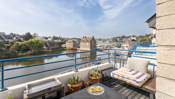 En balkon med udsigt over en flod, plantekasser, et bord med kop og underkop, samt en seng med pude med striber.
