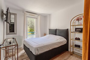 Quarto com cama, televisão na parede, mesa de vidro, prateleira com objetos e janela com cortinas.