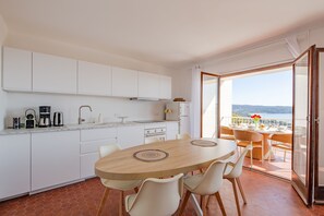 Una cocina moderna con armarios blancos, una mesa de comedor con sillas y vista al mar a través de un ventanal.
