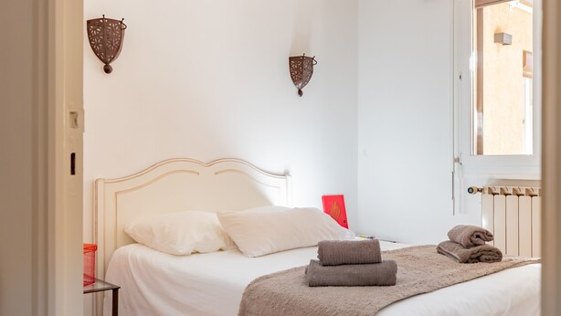 Ein Schlafzimmer mit Bett, Kissen, Decke, einem roten Gegenstand an der Wand und einem Fenster.