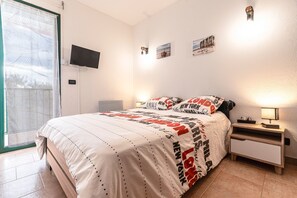 Un dormitorio con cama, televisor, mesita de noche y ventana con cortinas.