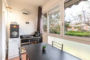 Una habitación compacta con una mesa de comedor, una planta pequeña en maceta, una cafetera, un altavoz y una ventana con vistas a zonas verdes y edificios.
