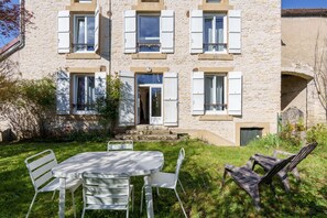 Ein Steinhaus mit weißen Fensterläden, einer Haustür und einem kleinen Garten mit Tisch und Stühlen.
