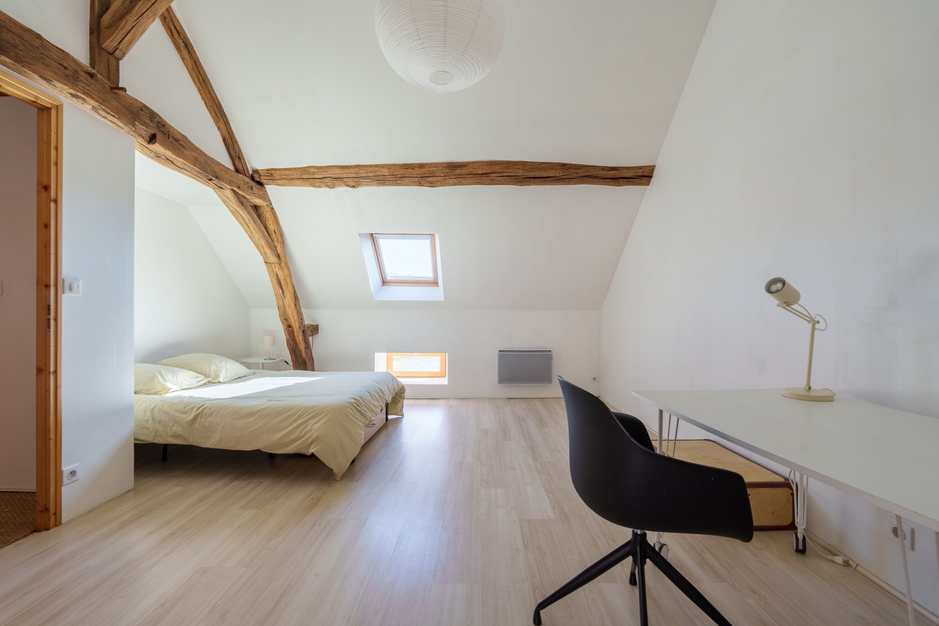 A room with a bed, desk, chair, and a sloped ceiling with exposed beams.