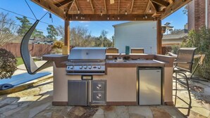 Een buitenkeuken met een barbecue, spoelbak en een inbouwkoelkast, onder een houten pergola met een plafondventilator.