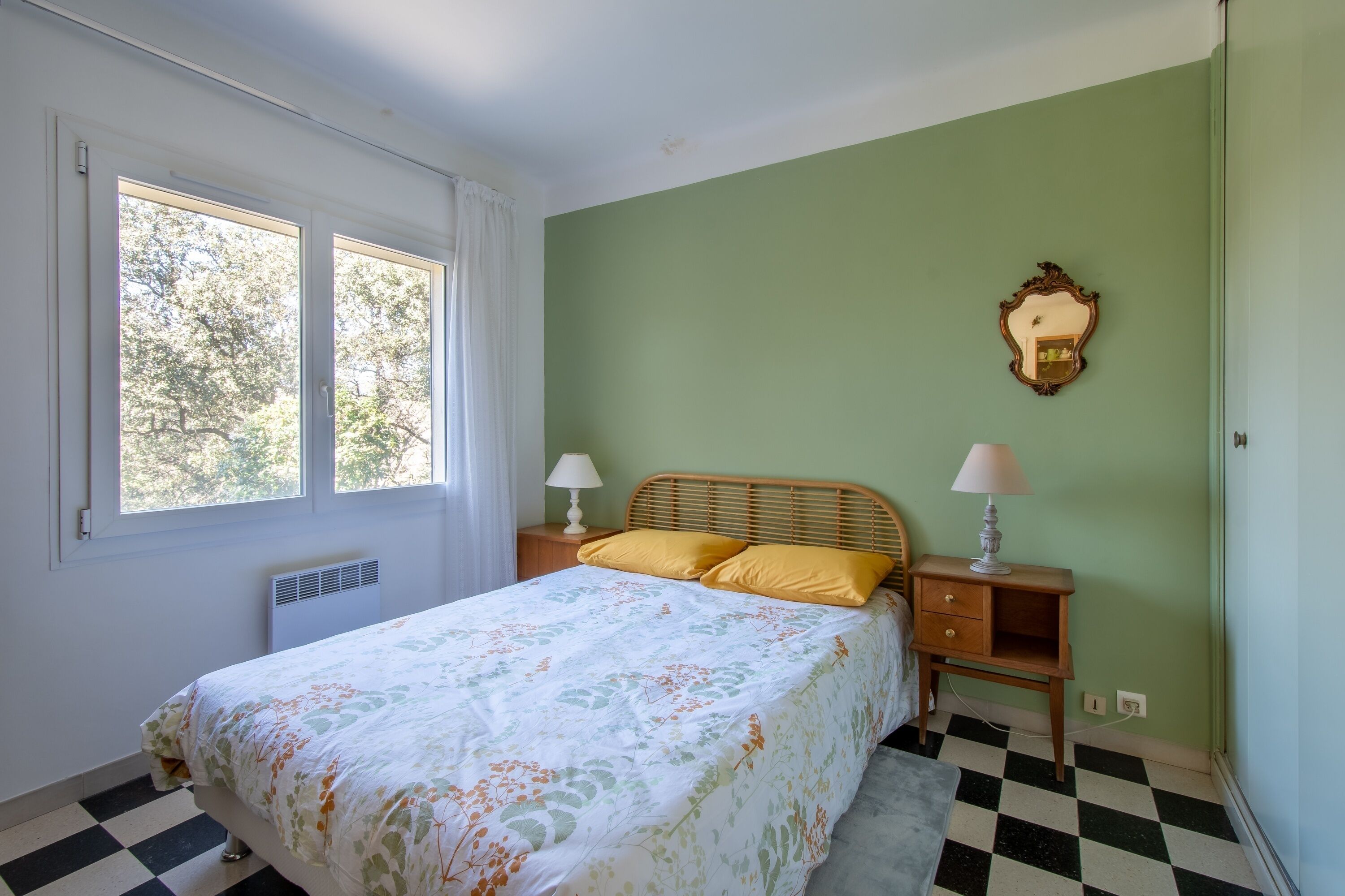 Une chambre à coucher avec un lit, deux tables de chevet avec des lampes, un radiateur, une fenêtre et un sol à carreaux.