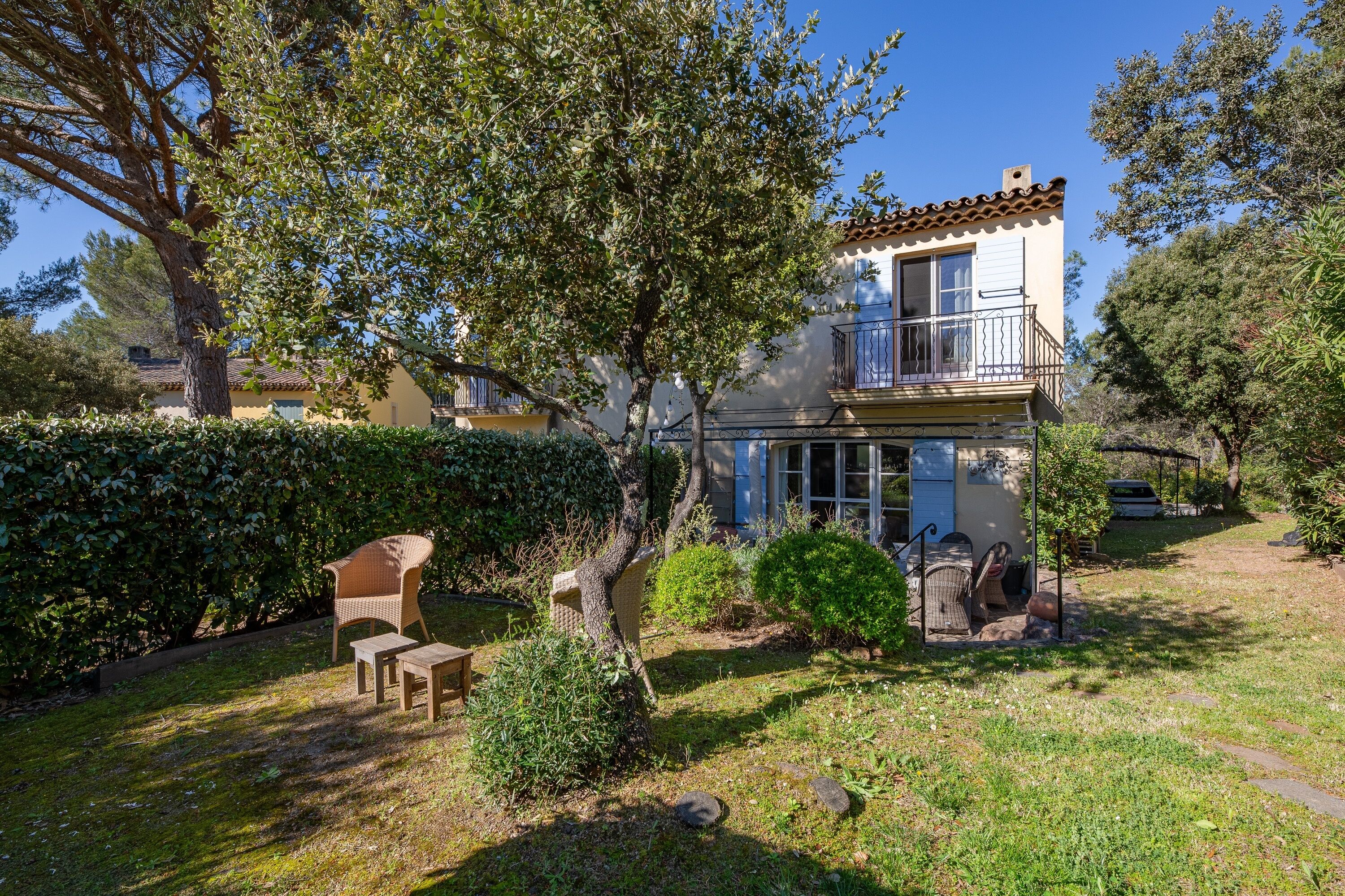 Une maison à deux étages avec un balcon, un jardin avec une chaise et une table, et un arbre au premier plan.