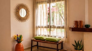 Ein Zimmer mit einer Bank, einem Spiegel, einer Blumenvase, einem Fenster mit transparenten Vorhängen und einem Regal mit Dekoration.
