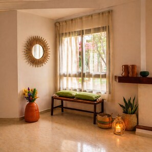 Ein Zimmer mit einer Bank, einem Spiegel, einer Blumenvase, einem Fenster mit transparenten Vorhängen und einem Regal mit Dekoration.