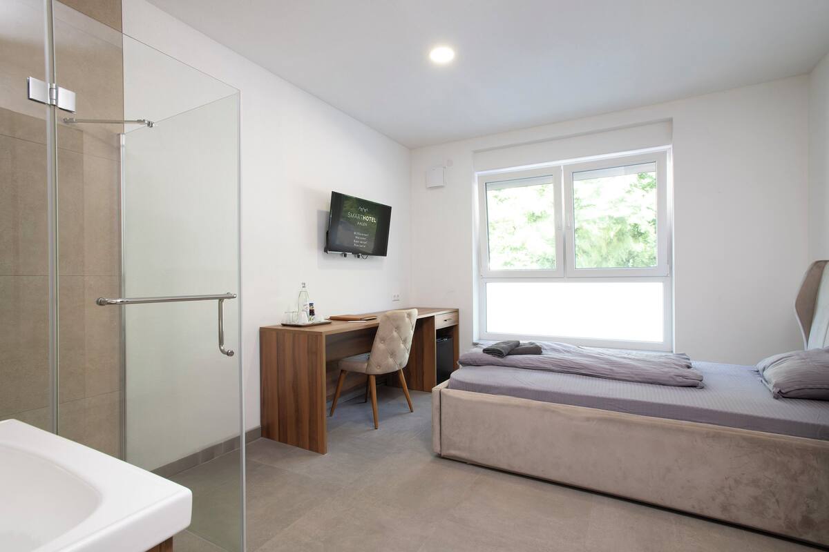 A modern hotel room with a shower, a desk, a bed, and a TV mounted on the wall.